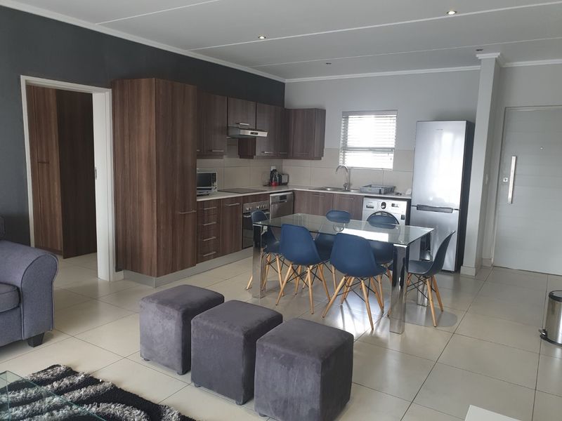 Open-plan kitchen and dining area featuring modern wood-finish cabinetry, integrated appliances, tiled flooring, and recessed lighting in a contemporary apartment setting.