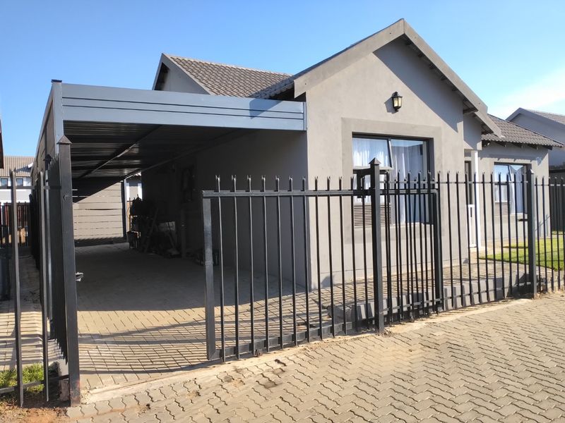 Front view of a townhouse featuring a paved driveway, covered carport, and secure metal perimeter fencing.