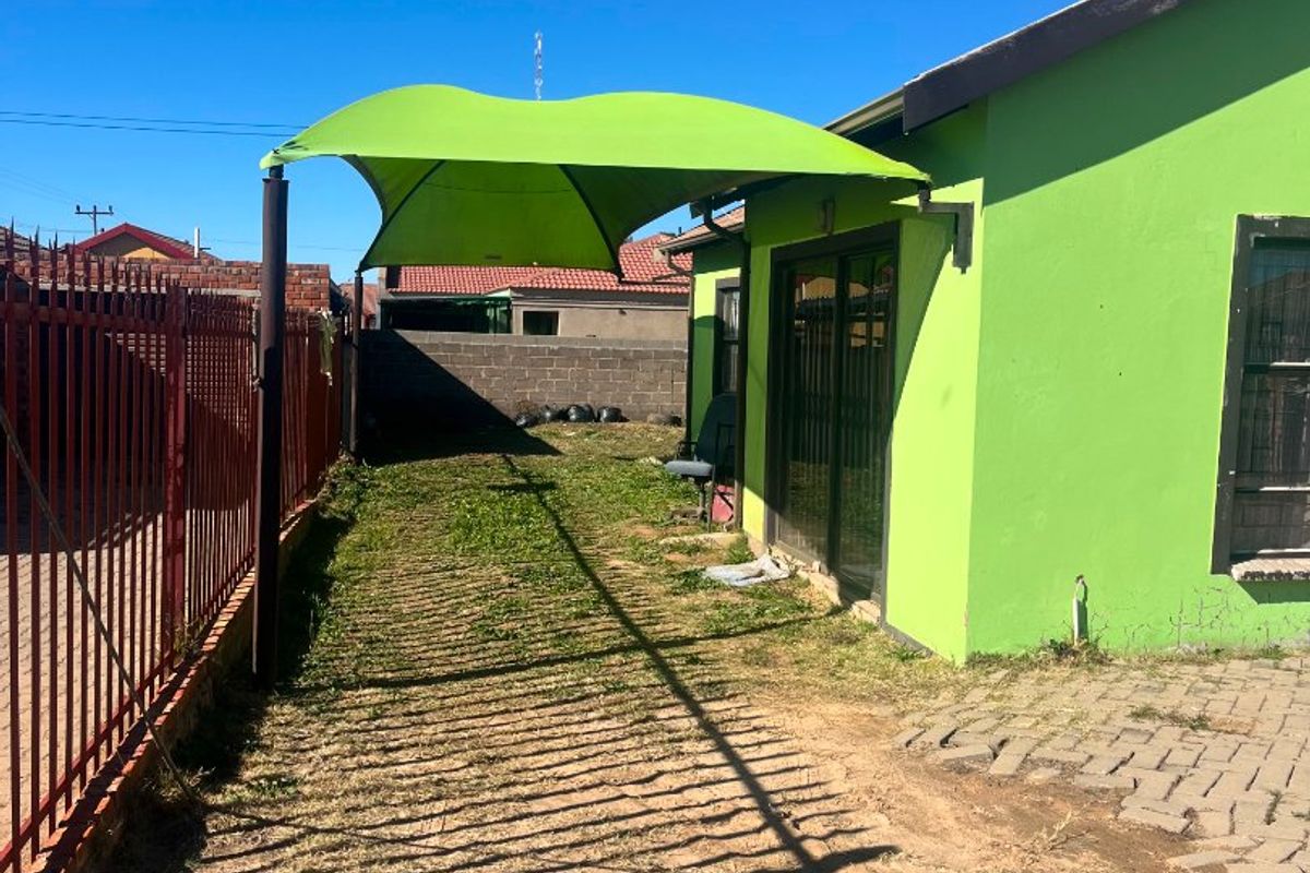 Rear exterior view featuring a green shade sail, sliding glass doors, and a metal perimeter fence with a paved walkway.
