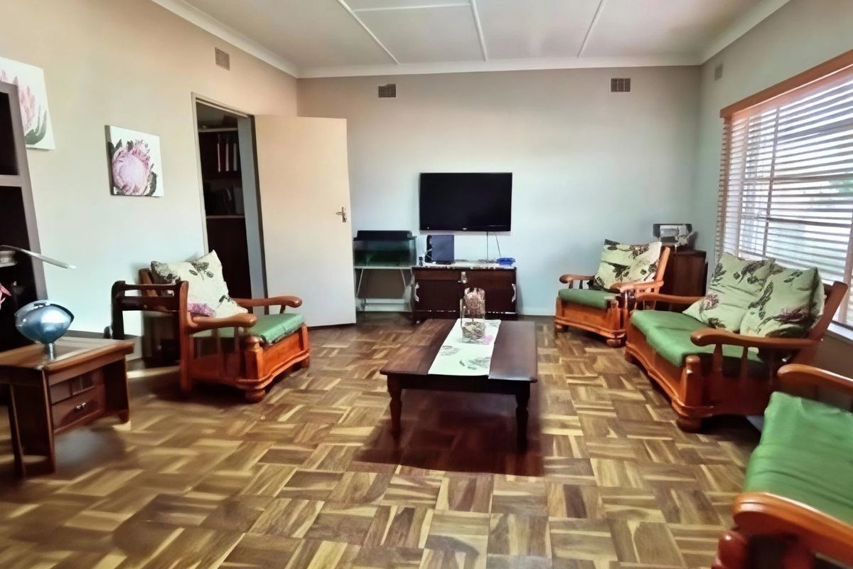 A spacious living room featuring classic parquet flooring, neutral wall tones, and large windows fitted with venetian blinds.