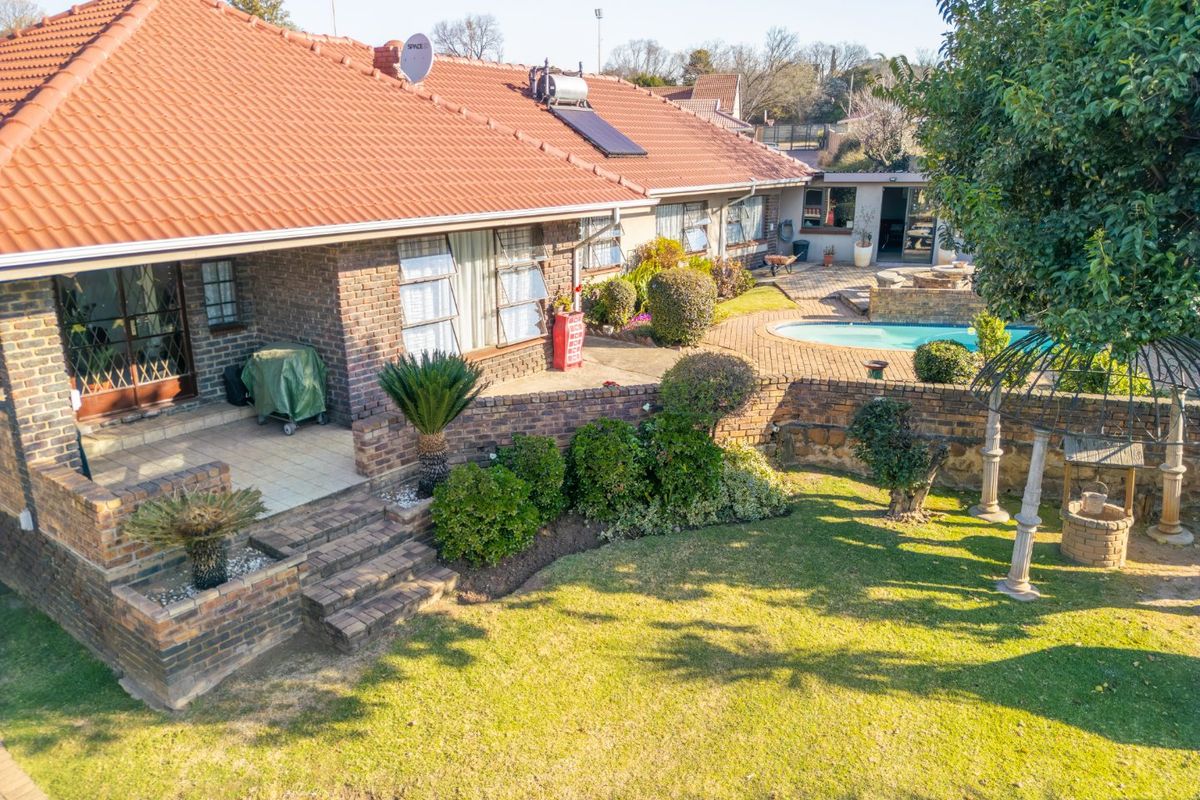 Aerial view of the rear exterior featuring a brick house, tiled roof, swimming pool, covered patio, and a well-maintained garden with brick retaining walls.