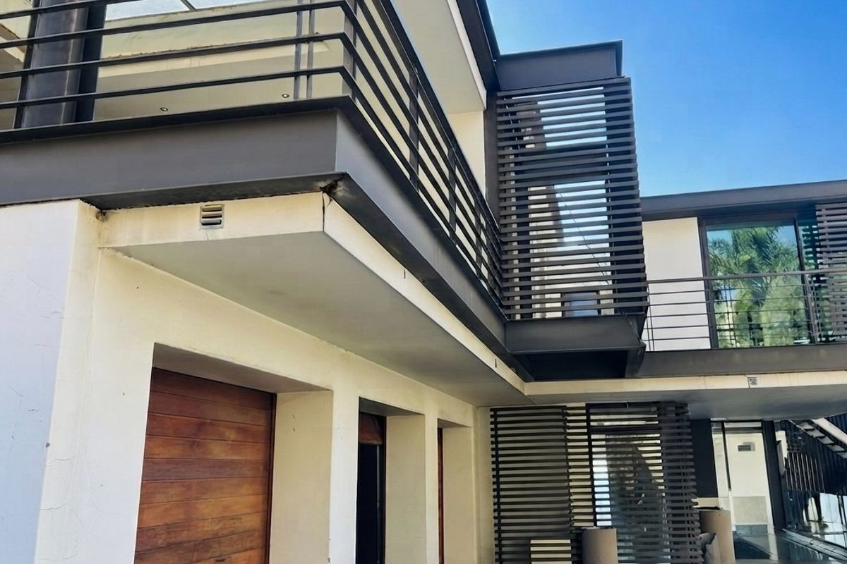 Modern architectural rear elevation featuring a paved courtyard, timber garage door, and upper-level balcony with metal railings and privacy screens.