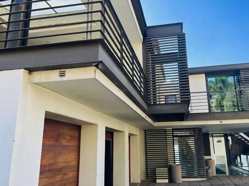 Modern architectural rear elevation featuring a paved courtyard, timber garage door, and upper-level balcony with metal railings and privacy screens.