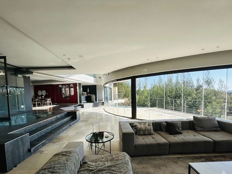 Spacious open-plan living area featuring expansive curved glass walls, stone flooring, and a sunken lounge design with seamless indoor-outdoor flow.