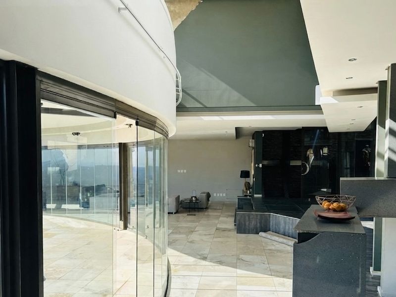 Spacious open-plan living area featuring a striking curved glass wall, stone flooring, and a raised platform, offering expansive views and modern architectural design.