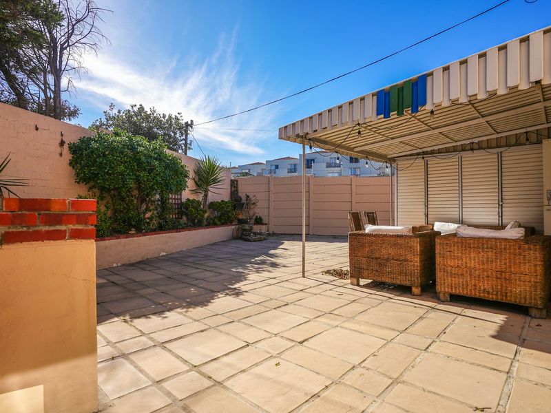 A paved outdoor patio area featuring a fixed metal awning, brick retaining wall, and perimeter boundary walls, offering a functional space for outdoor living.