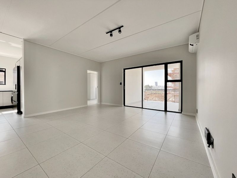 A bright, modern open-plan living area featuring neutral tiled floors, track lighting, and direct access to a private balcony through large sliding glass doors.