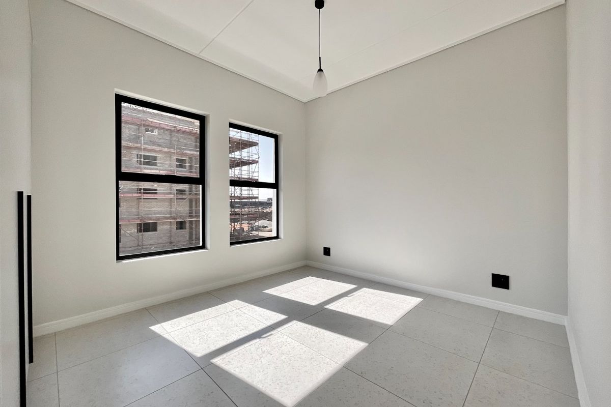 A bright, modern, and empty bedroom featuring neutral tiled flooring, clean white walls, and large black-framed windows allowing for ample natural light.