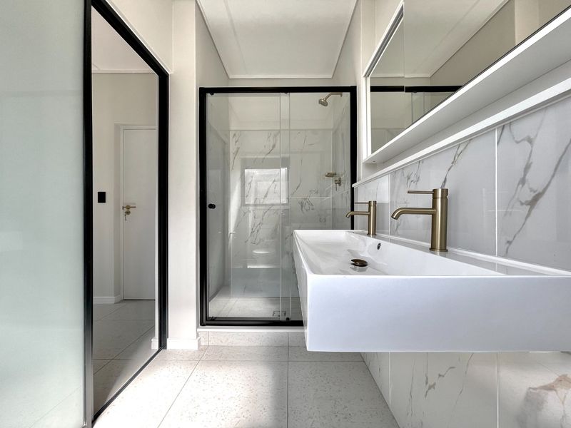 Modern bathroom featuring a floating double vanity with brass fixtures, marble-look wall tiles, and a spacious walk-in shower with a glass enclosure.