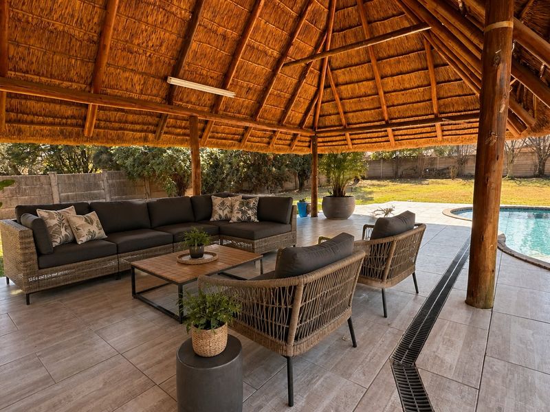 A spacious covered patio featuring a traditional thatched roof, timber support beams, and tiled flooring, overlooking a private swimming pool and garden area.