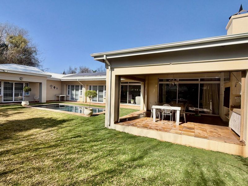 Spacious rear exterior featuring a covered patio, tiled flooring, built-in outdoor cabinetry, and a swimming pool set within a well-maintained garden.
