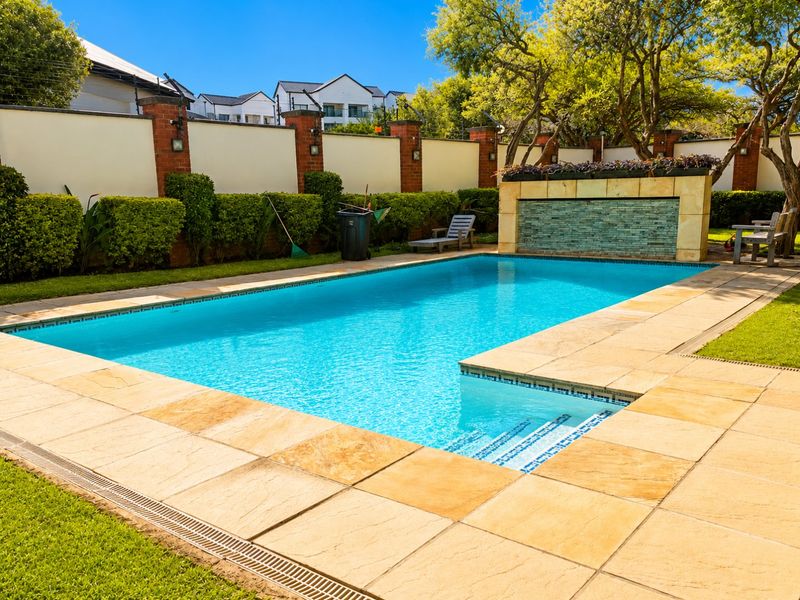 A well-maintained swimming pool area featuring stone paving, a decorative water feature, and secure perimeter walling in a landscaped garden setting.