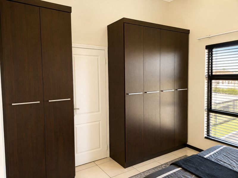A well-maintained bedroom featuring dark wood built-in cupboards, neutral tiled flooring, and a window fitted with venetian blinds.