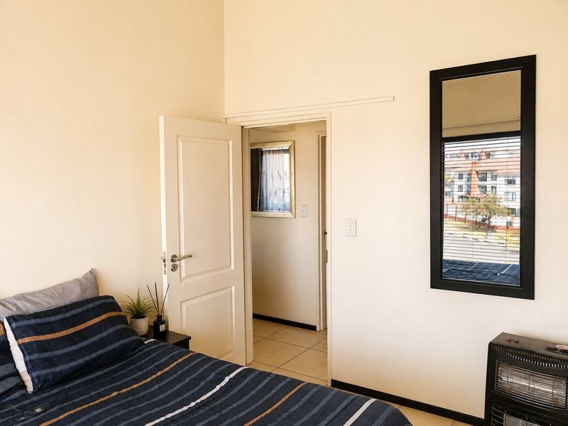 A bedroom featuring tiled flooring, a neutral wall finish, and a window with a black frame, providing access to an adjacent hallway.