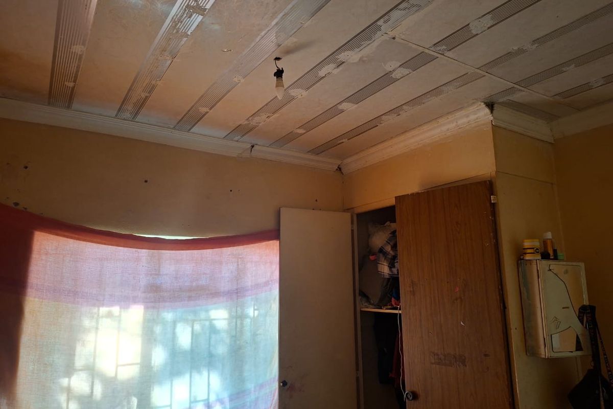 A bedroom featuring a built-in wardrobe, security bars on the window, and a tiled ceiling. The room requires significant cosmetic updates and repairs.