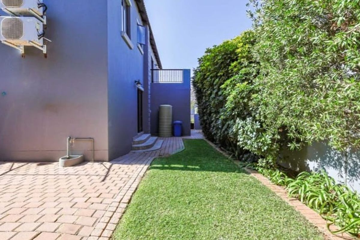 A well-maintained rear garden featuring a paved walkway, manicured lawn, boundary hedge, and exterior wall-mounted air conditioning units.