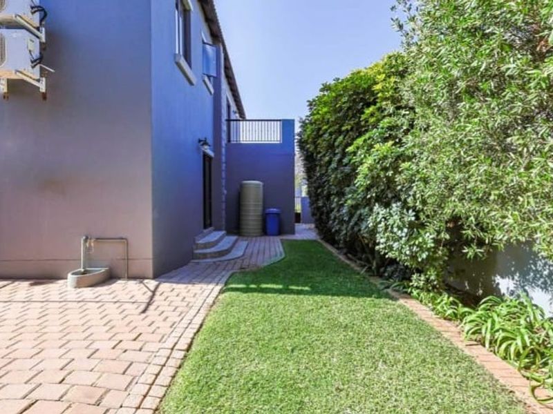 A well-maintained rear garden featuring a paved walkway, manicured lawn, boundary hedge, and exterior wall-mounted air conditioning units.