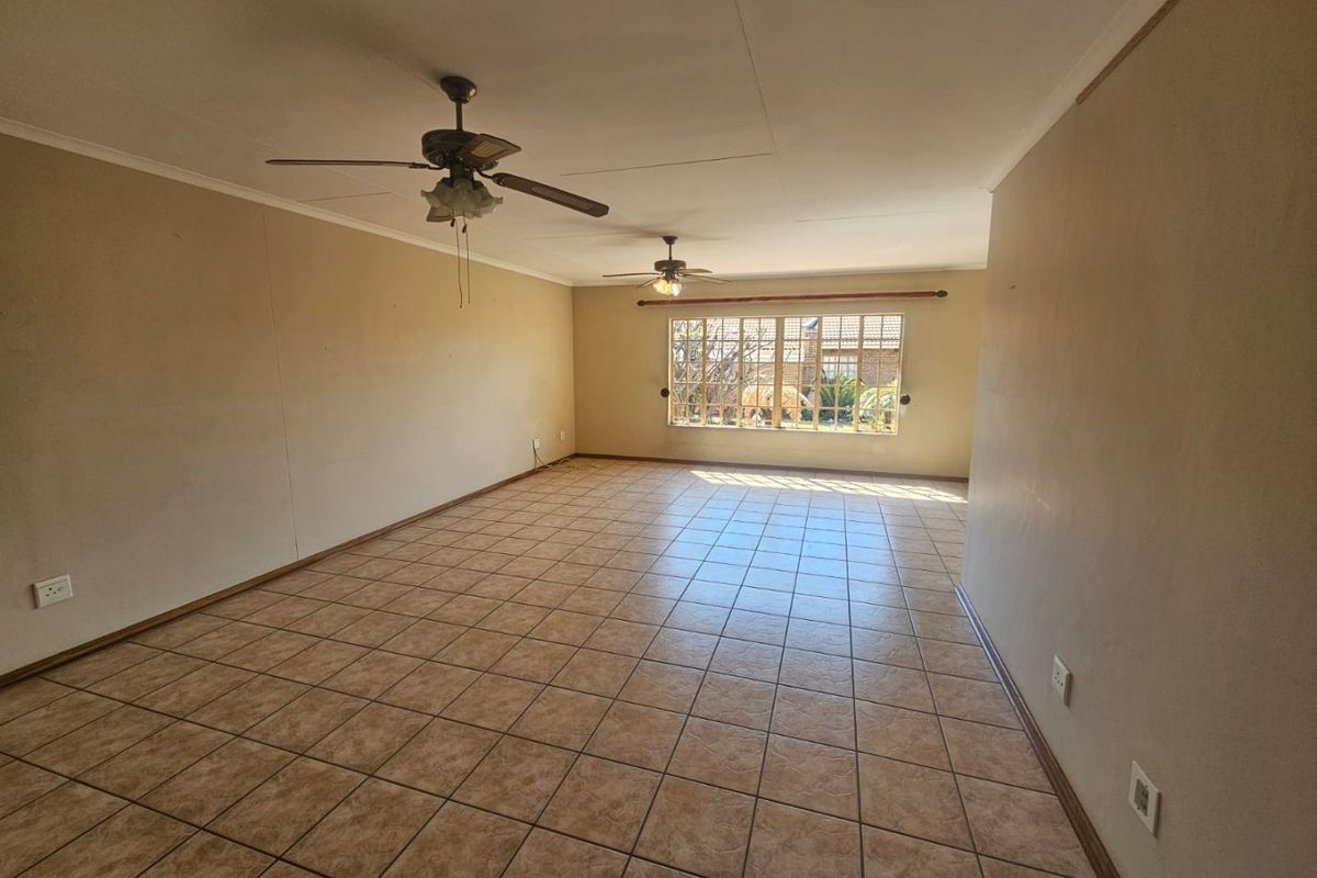 Spacious, empty living area featuring neutral tiled flooring, dual ceiling fans, and a large window providing natural light.