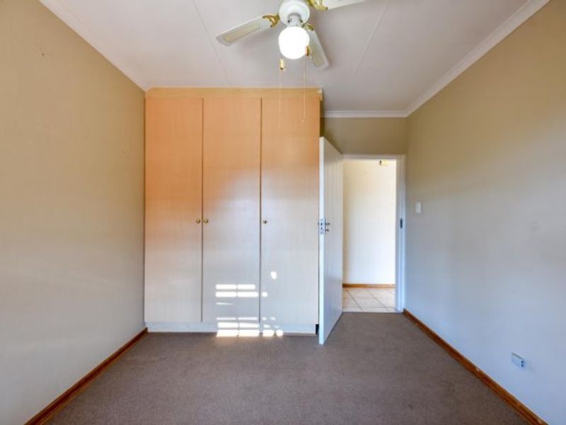 A clean, empty bedroom featuring built-in wooden cupboards, neutral wall finishes, carpeted flooring, and a central ceiling fan.
