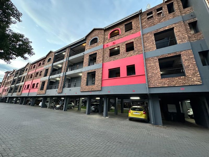 Exterior view of a multi-story apartment building featuring a brick facade, private balconies, and ground-level covered parking bays.