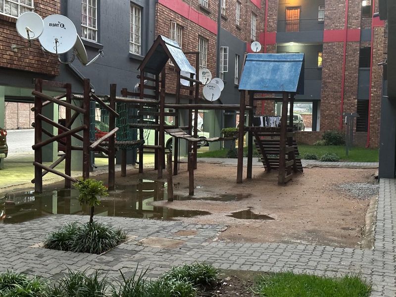 Communal outdoor area featuring a playground structure, paved pathways, and residential building facades with satellite dish installations.