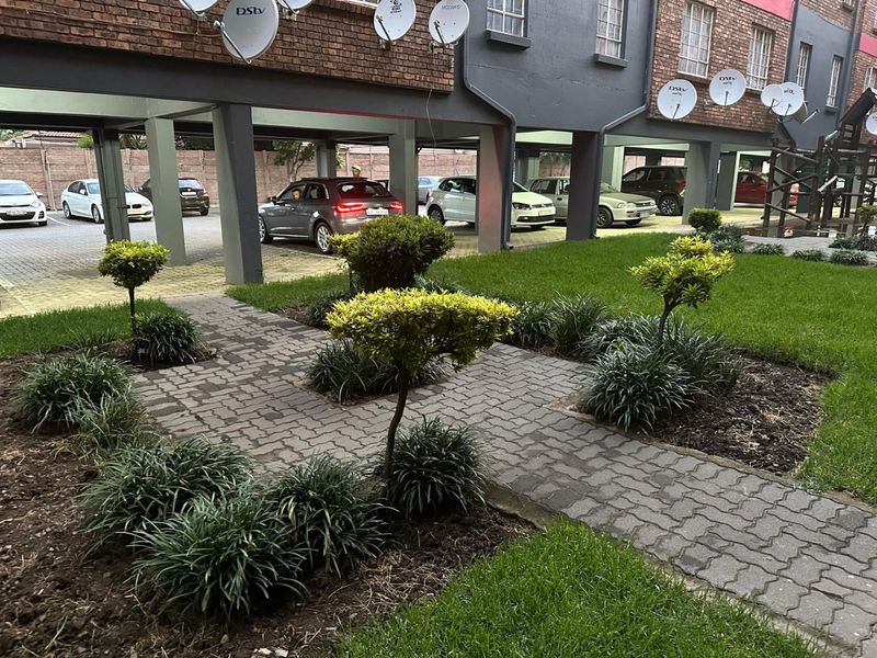 A view of the apartment complex exterior featuring a landscaped garden, paved walkway, and a covered parking area with concrete support pillars.