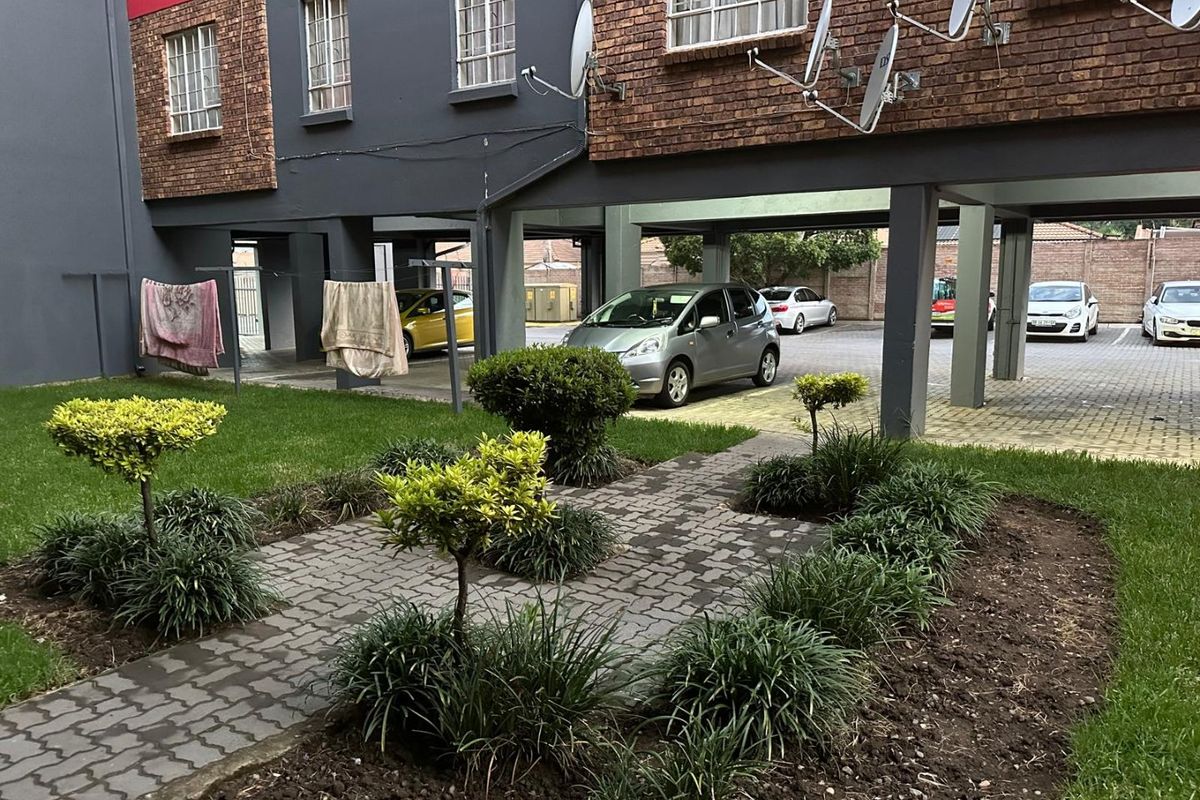 A view of the apartment complex rear exterior featuring a landscaped garden, paved walkway, and covered parking area with brick facade detailing.