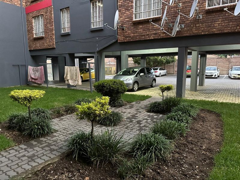 A view of the apartment complex rear exterior featuring a landscaped garden, paved walkway, and covered parking area with brick facade detailing.