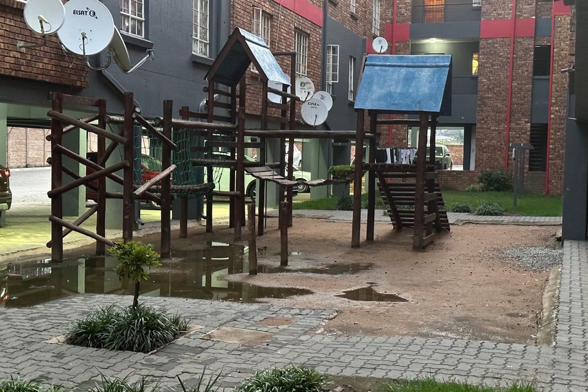 Communal outdoor area featuring a playground structure, paved pathways, and residential building facades with satellite dish installations.