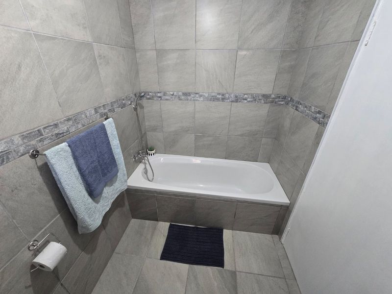 A modern bathroom featuring a built-in bathtub with wall-mounted shower mixer, finished with neutral grey ceramic wall and floor tiles.