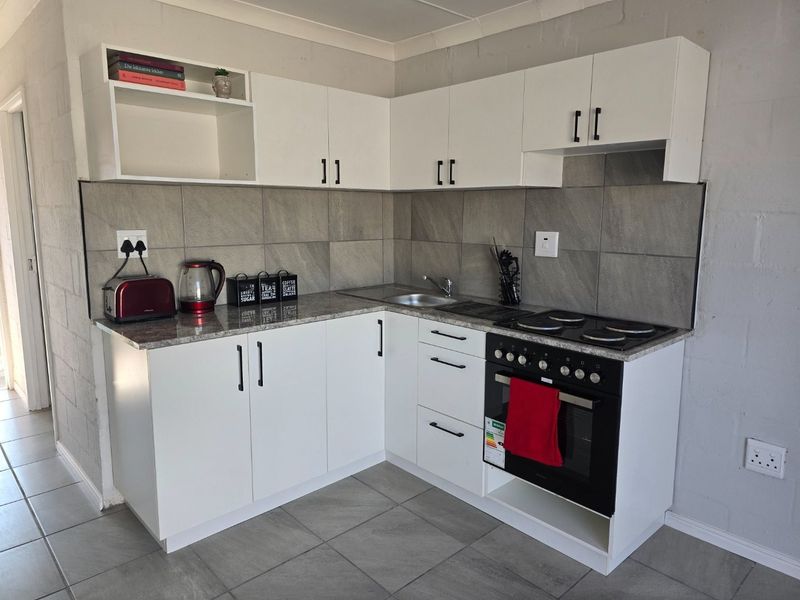 A compact, modern kitchen featuring white cabinetry, granite countertops, a built-in oven with electric hob, and neutral tiled backsplash and flooring.
