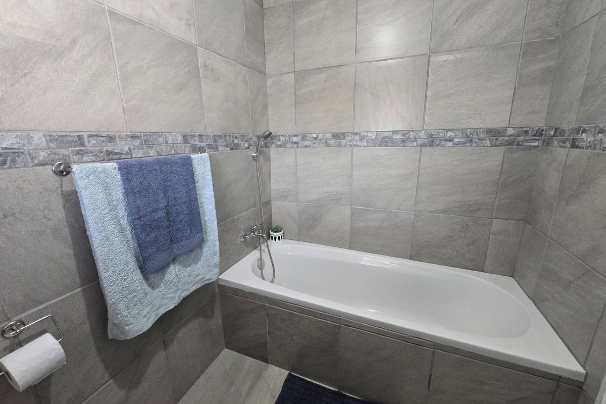 A modern bathroom featuring a built-in bathtub with a handheld shower mixer, finished with neutral grey ceramic wall tiles.