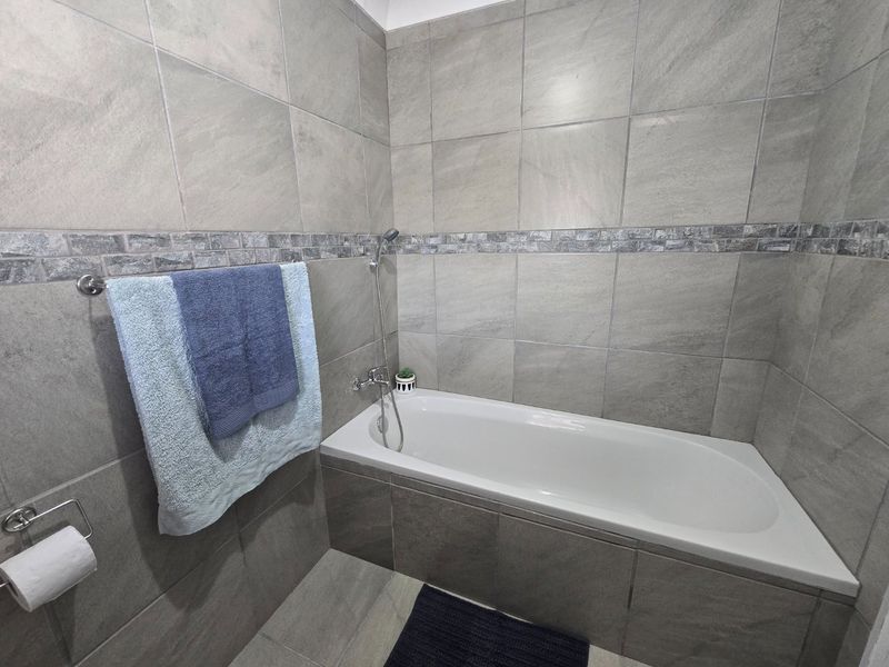 A modern bathroom featuring a built-in bathtub with a handheld shower mixer, finished with neutral grey ceramic wall tiles.
