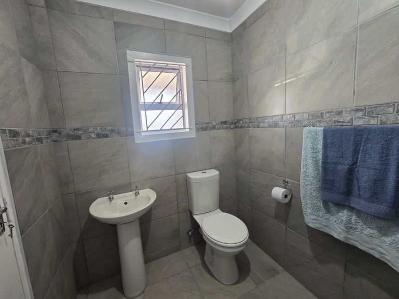 A modern, fully tiled bathroom featuring a pedestal basin and a standard toilet, finished with neutral grey wall and floor tiles.