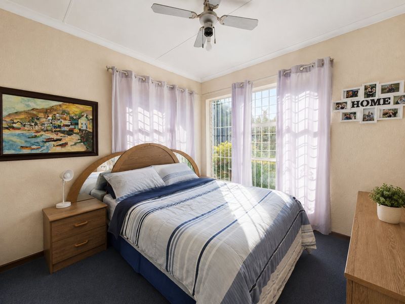 A well-lit bedroom featuring carpet flooring, a ceiling fan, and large windows with security bars, offering a comfortable and functional living space.