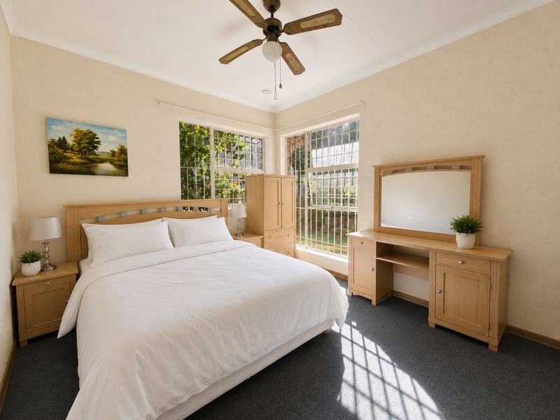 A well-lit bedroom featuring carpeted flooring, a ceiling fan, and large windows with security bars, offering a comfortable and functional living space.