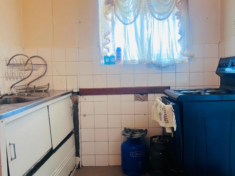 A dated kitchen featuring a stainless steel sink, tiled splashback, and freestanding stove, currently showing significant wear and missing wall tiles.