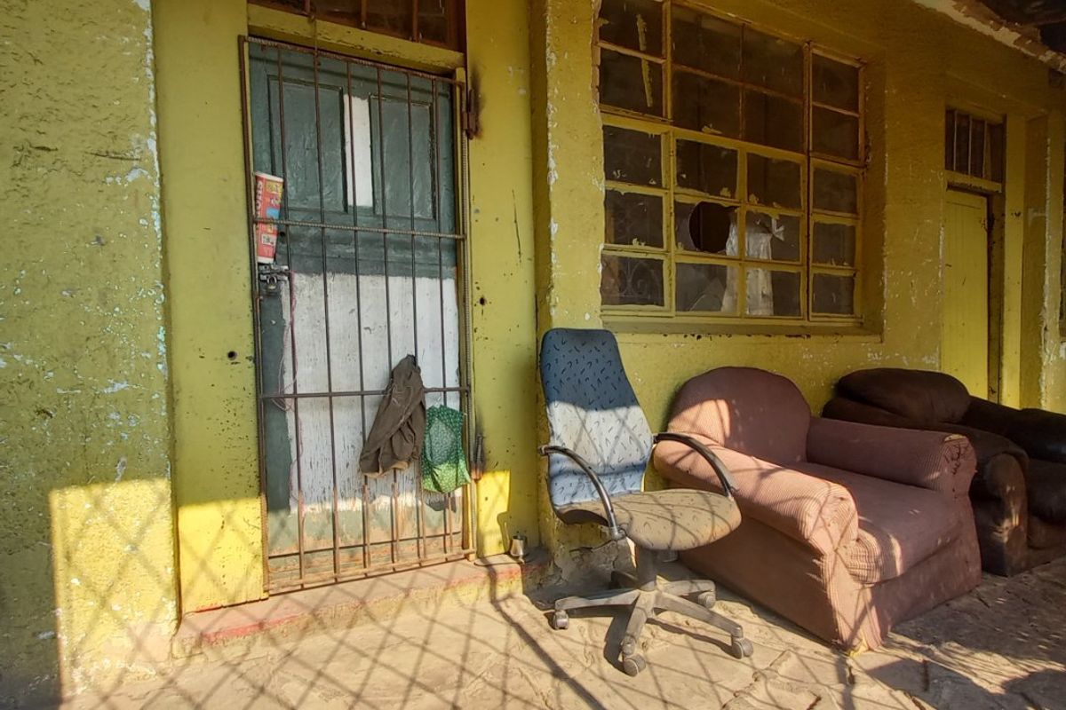 A covered patio area featuring stone paving, steel-framed windows, and a security gate, currently requiring significant maintenance and exterior restoration.