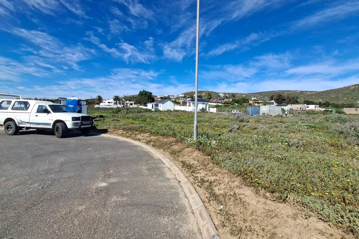 A view of a vacant residential plot from the adjacent paved road, featuring natural vegetation and surrounding neighborhood development.