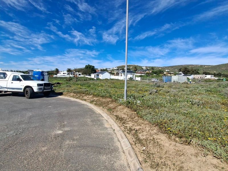 A view of a vacant residential plot from the adjacent paved road, featuring natural vegetation and surrounding neighborhood development.