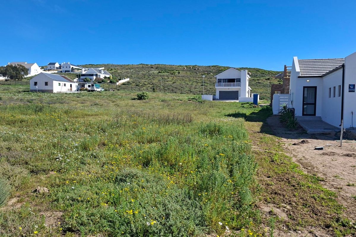 A view of a vacant residential plot with natural vegetation, adjacent to existing modern homes and paved driveway access.
