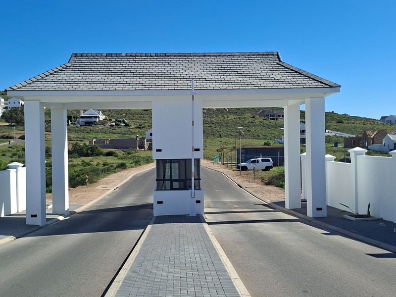 A well-maintained residential estate entrance featuring a modern security gatehouse, paved driveway, and perimeter boundary walls under a clear sky.