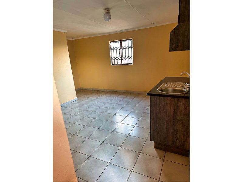 A compact, empty kitchen area featuring tiled flooring, basic cabinetry with a stainless steel sink, and a window with security bars.