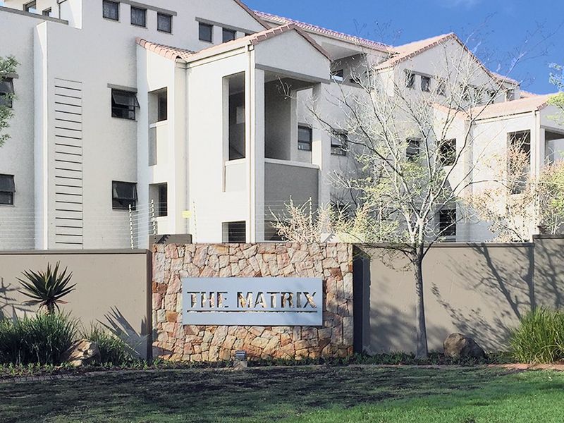 Front elevation of The Matrix apartment complex featuring a stone-clad entrance wall, perimeter security fencing, and multi-story residential building structure.