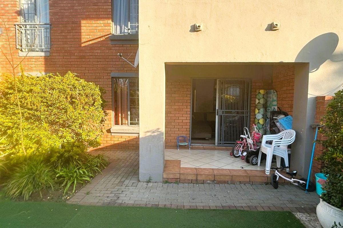 Rear exterior view of the apartment featuring a covered tiled patio, brick facade, and access to a small private garden area with artificial lawn.