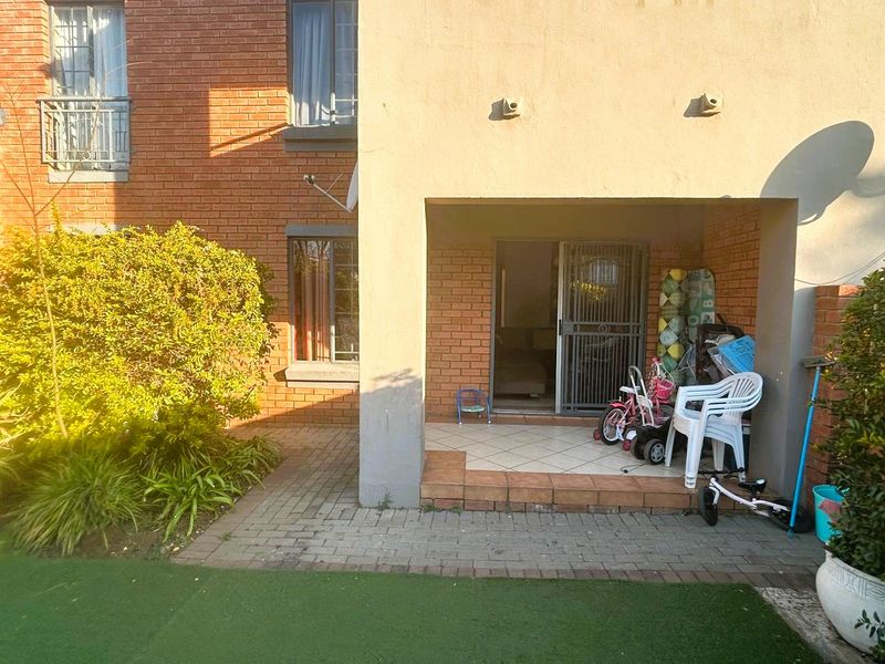 Rear exterior view of the apartment featuring a covered tiled patio, brick facade, and access to a small private garden area with artificial lawn.