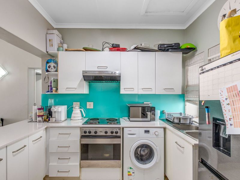 Compact kitchen featuring white cabinetry, a vibrant turquoise glass splashback, integrated washing machine, and built-in electric oven with cooktop.