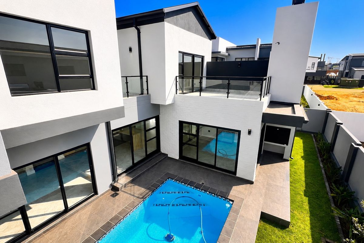 Modern rear exterior view featuring a swimming pool, tiled patio area, built-in braai, and an upper-level balcony with glass balustrades.