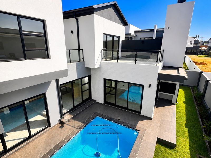 Modern rear exterior view featuring a swimming pool, tiled patio area, built-in braai, and an upper-level balcony with glass balustrades.