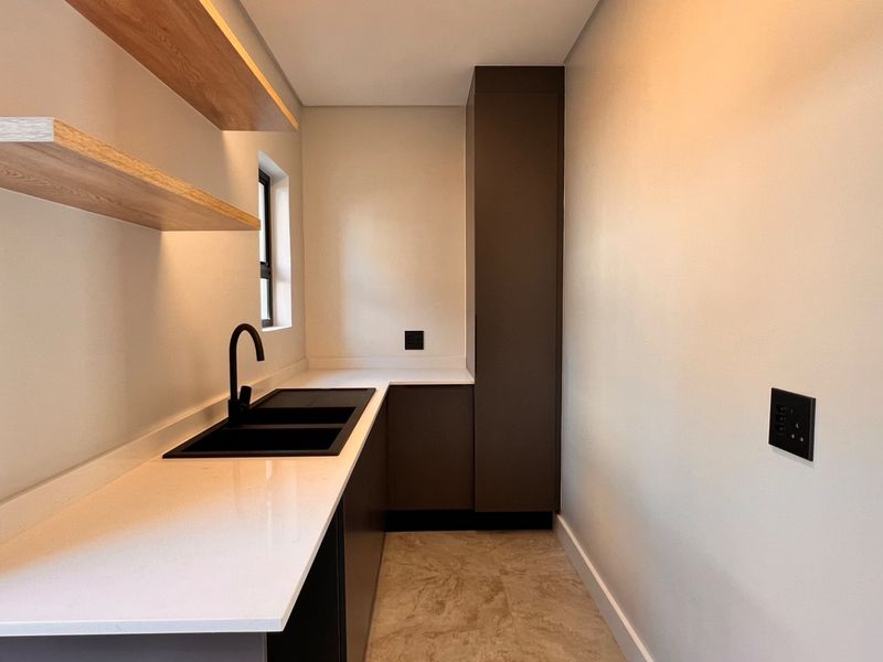 A modern, minimalist scullery featuring sleek dark cabinetry, white stone countertops, a black sink, and floating timber shelving.